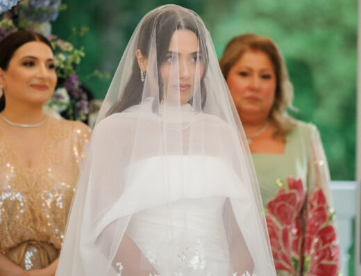 A bride in a white dress and veil stands with her hands clasped, flanked by two women in formal attire; floral decorations are visible in the background.