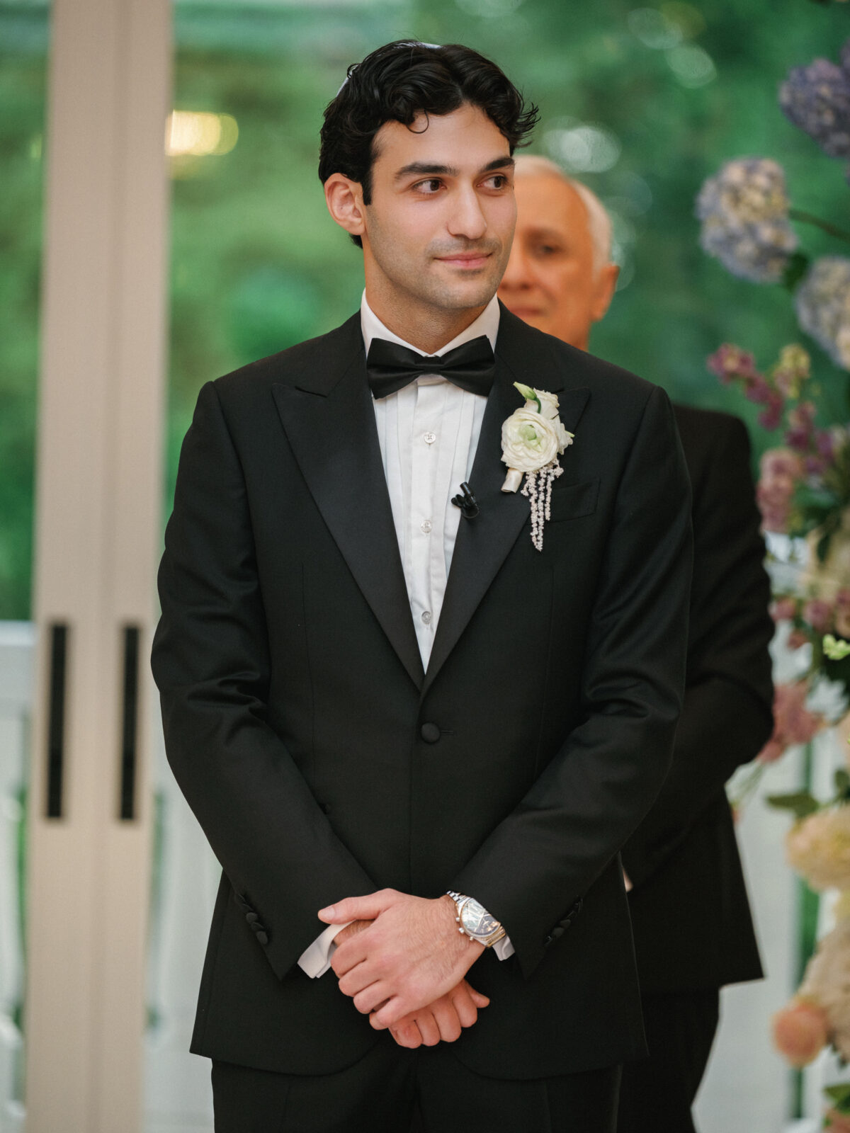 A man in a black tuxedo with a white boutonniere stands indoors, hands clasped, with greenery and floral decorations in the background.