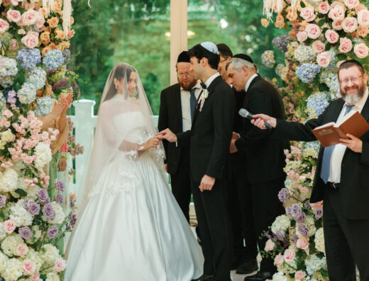A bride and groom exchange rings under a floral wedding canopy, surrounded by officiants and witnesses, during a ceremony.