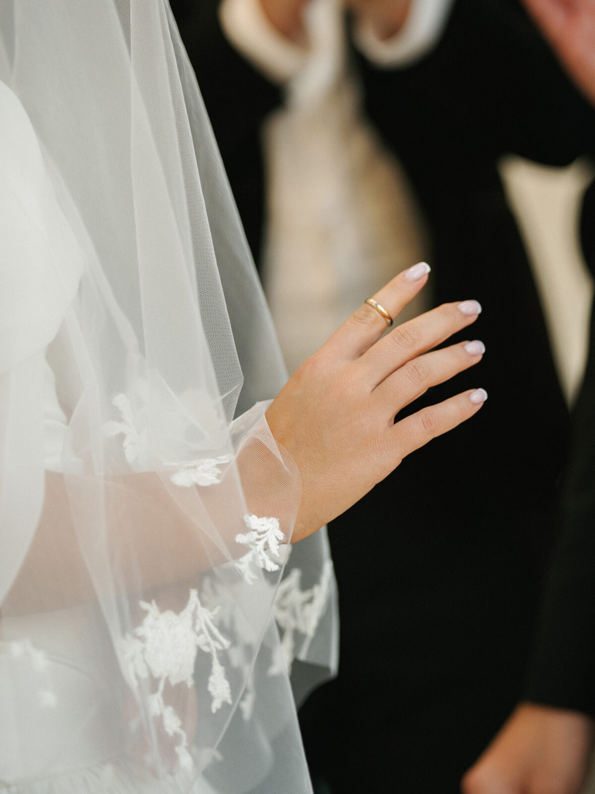 A close-up of a hand wearing a gold ring, partially covered by a white lace wedding veil, with blurred people in dark clothing in the background.