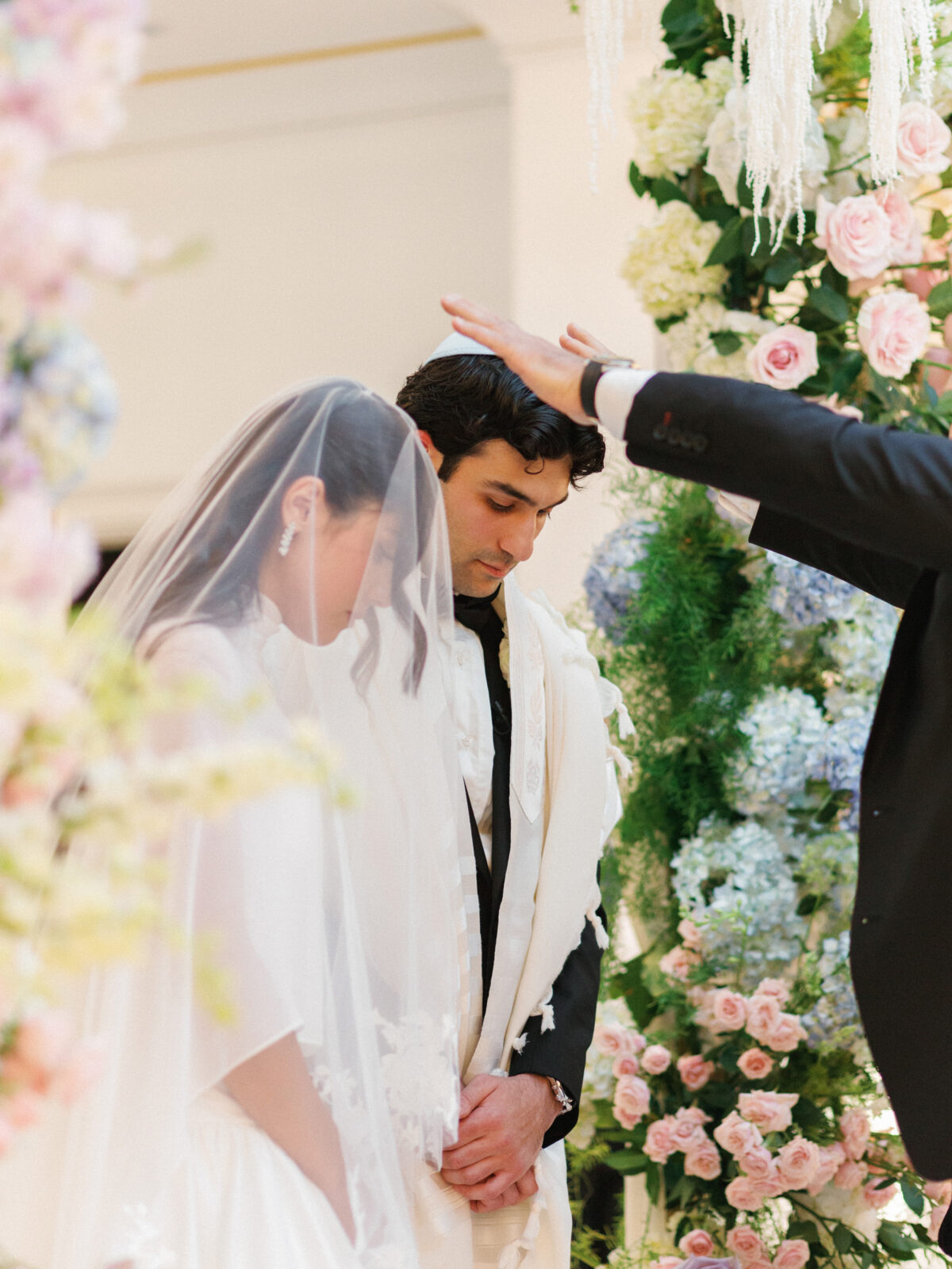 A bride and groom stand with heads bowed under a floral arch while an officiant raises a hand over them during a wedding ceremony.