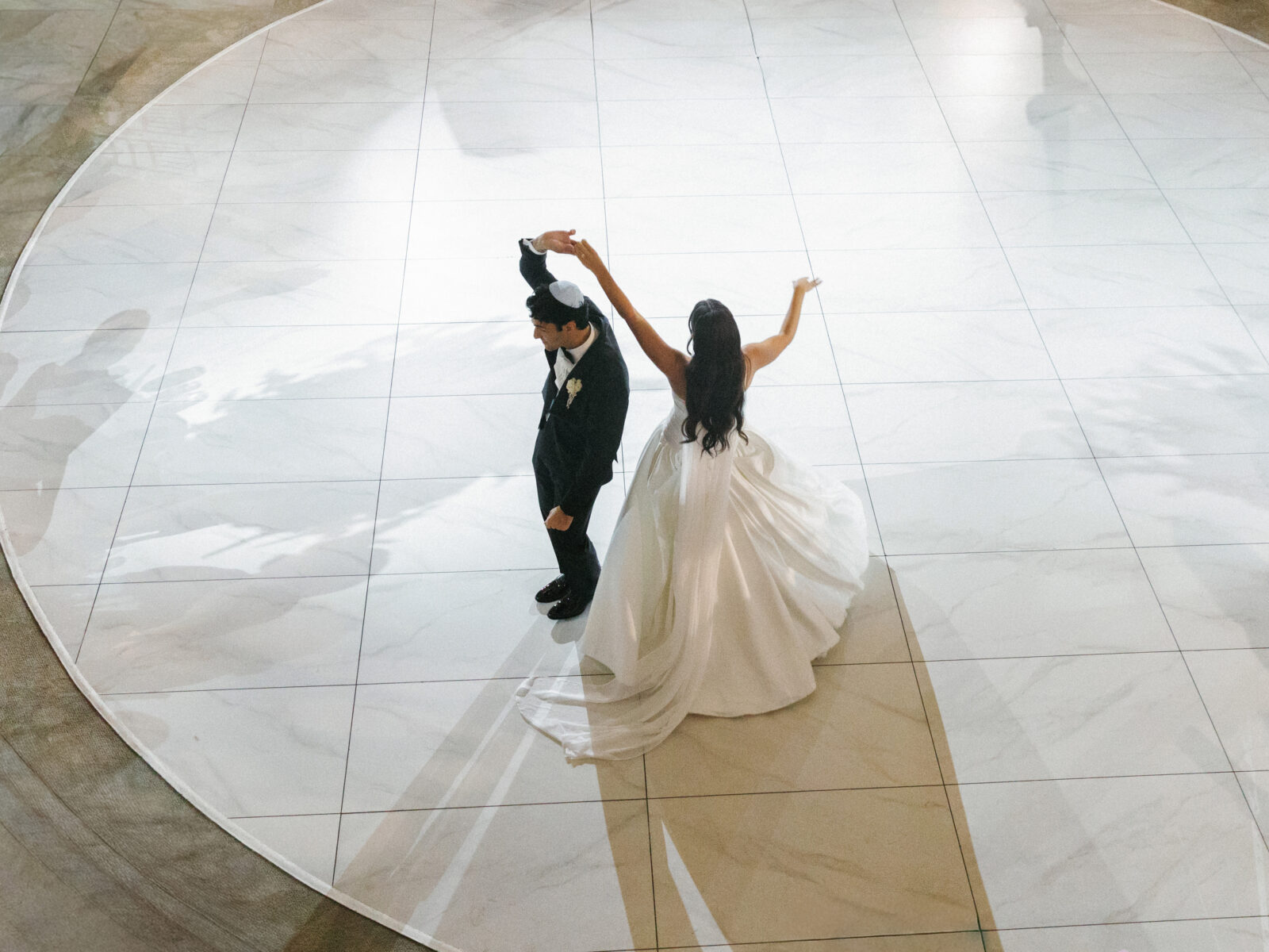 A bride and groom dance together on a large, circular white floor, with the bride holding up one arm and the groom in a black suit beside her.