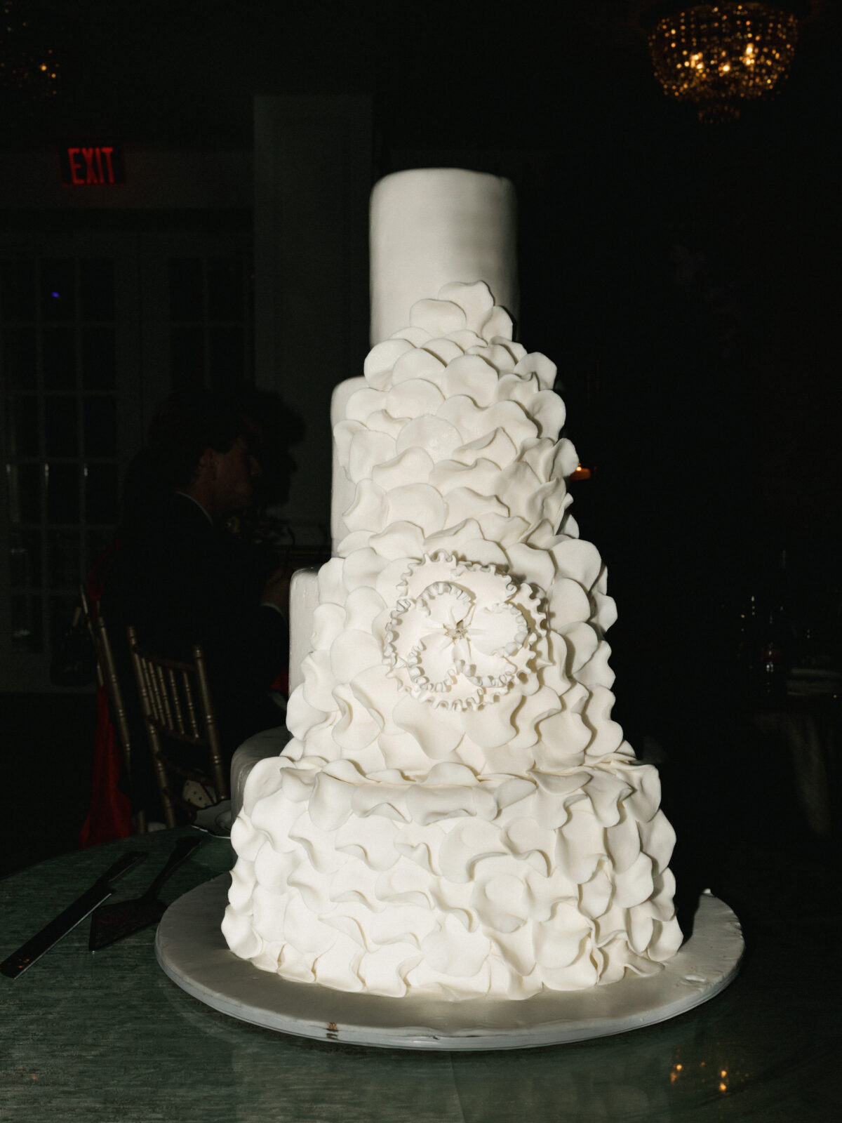 A tall, tiered white wedding cake decorated with ruffled icing and two interlocking rings in the center, displayed on a round table.