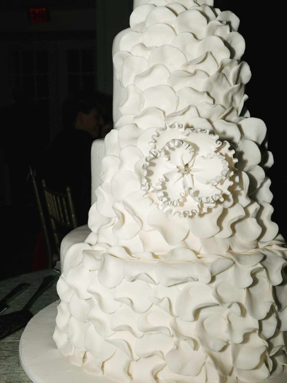 A tiered white cake decorated with ruffled icing and a floral design, displayed on a table at an indoor event.