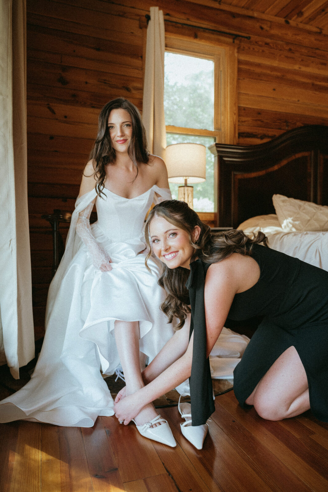 A bride in a white dress sits on a bed while another woman kneels and helps her put on white shoes in a wood-paneled room.