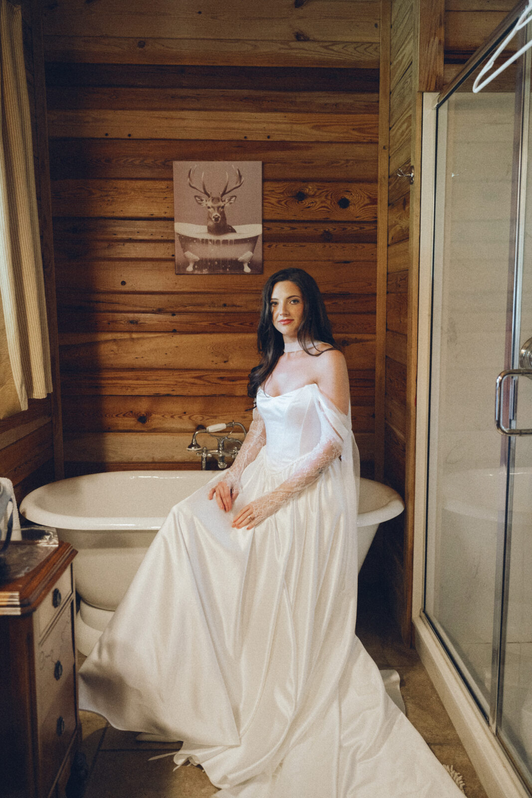A woman in a white wedding dress sits on the edge of a bathtub in a wooden bathroom, with a deer portrait hanging on the wall behind her.