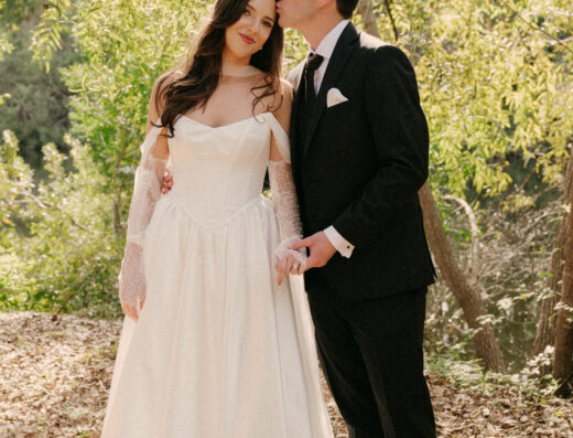 A bride in a white gown and a groom in a black suit stand together outdoors among trees, holding hands.