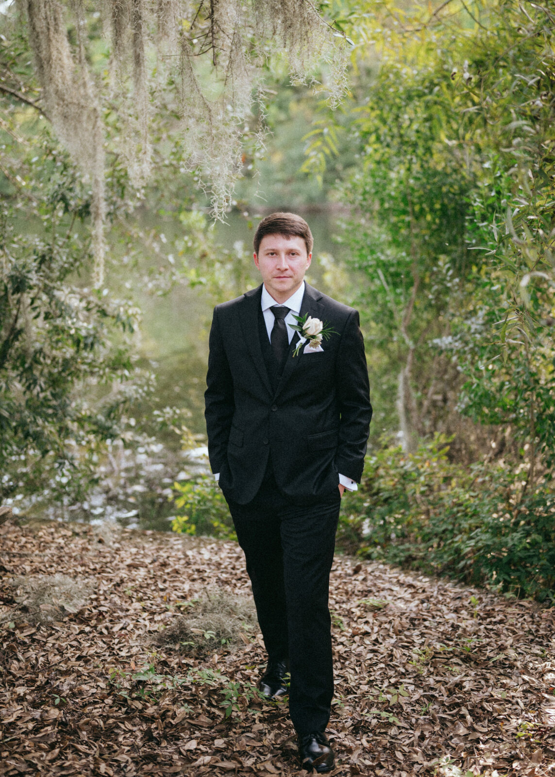 A man in a black suit with a boutonniere walks outdoors on a leaf-covered path, surrounded by green trees and moss, with a pond in the background.