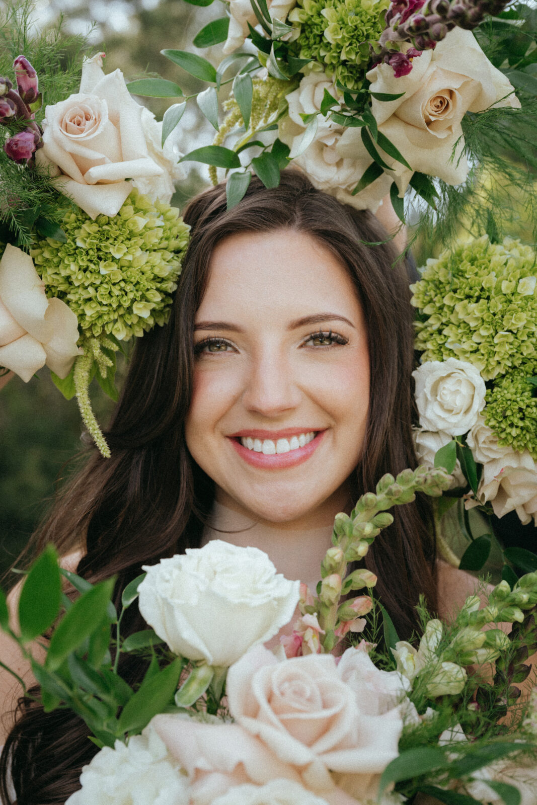 Woman with long brown hair smiles, surrounded by pale roses, green hydrangeas, and assorted greenery outdoors.