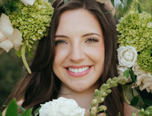 Woman with long brown hair smiles, surrounded by pale roses, green hydrangeas, and assorted greenery outdoors.