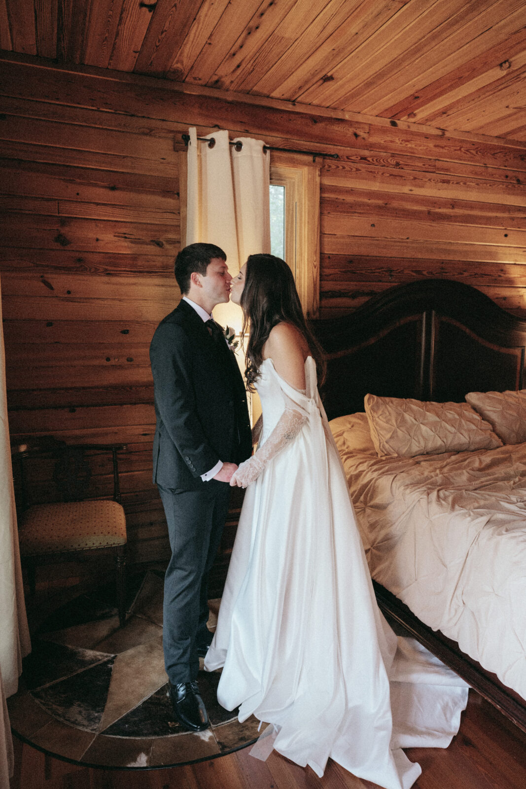 A bride and groom in formal attire stand face to face, holding hands, in a wooden-paneled bedroom with a made bed and curtained window.
