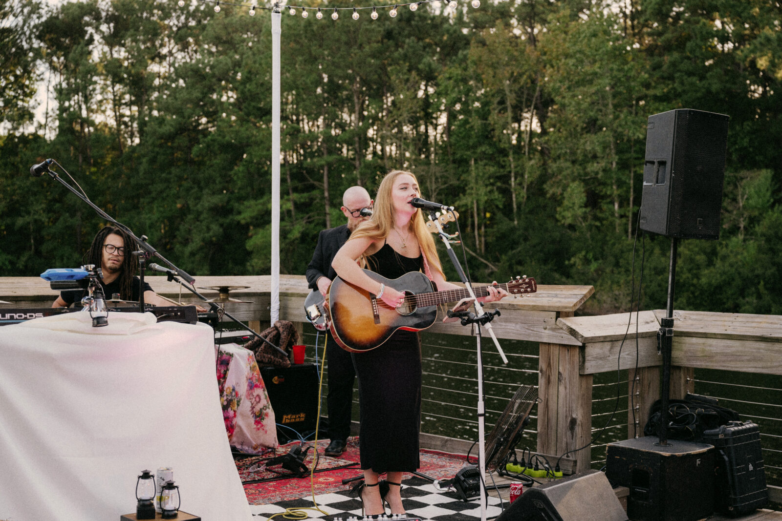 A woman sings and plays guitar on an outdoor stage with two musicians, one on keyboard and another on bass, in front of trees and string lights.