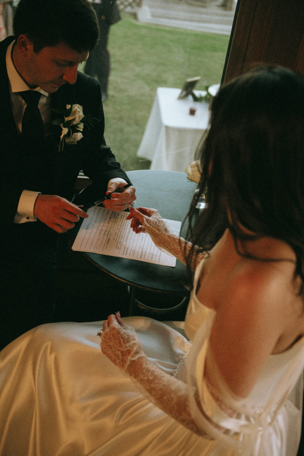 A bride and groom sit together indoors, signing a document on a table. The bride wears a white dress and lace gloves, and the groom is in a dark suit with a boutonniere.