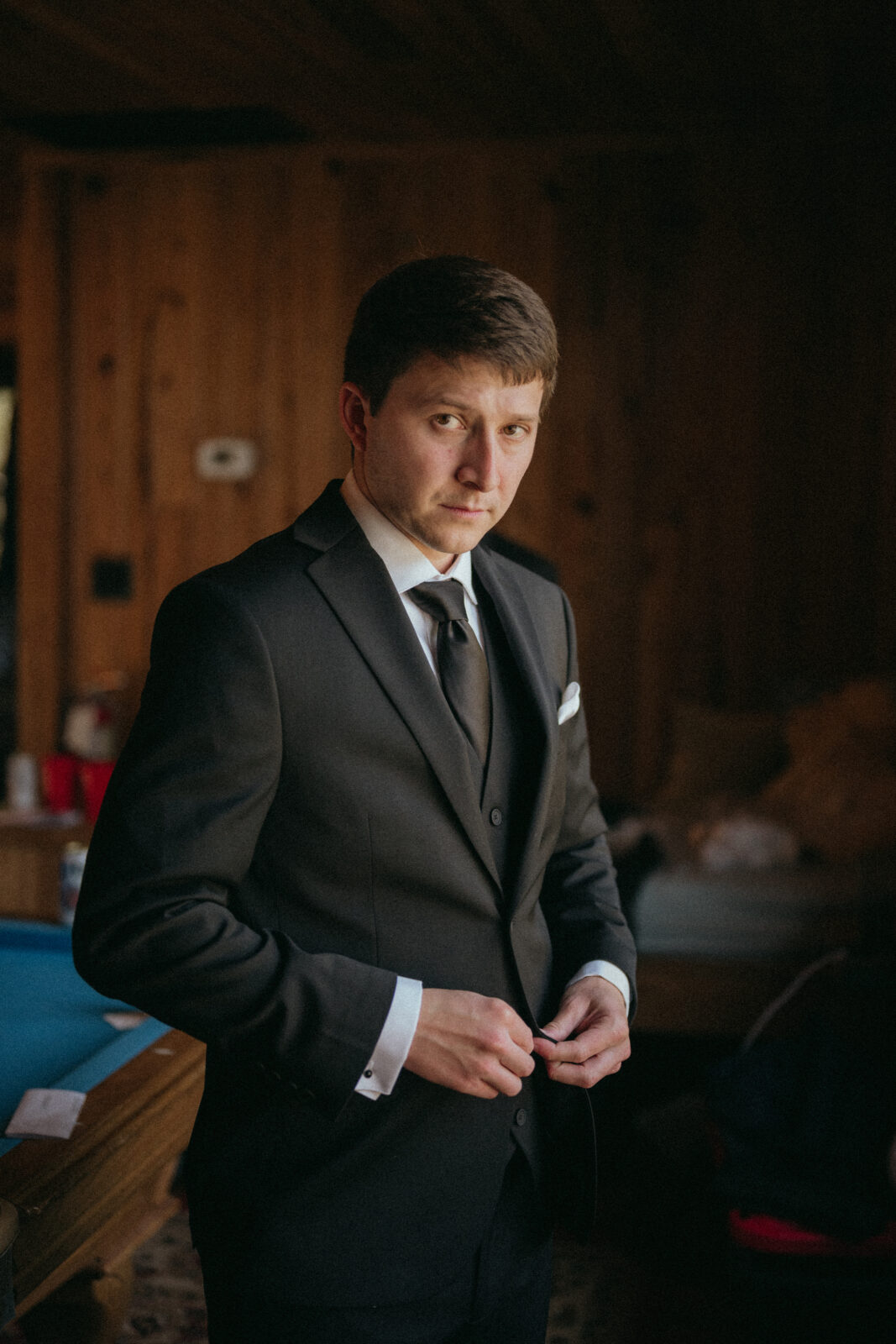 A man in a black suit stands indoors, buttoning his jacket and looking at the camera. The background features wood-paneled walls and a pool table.