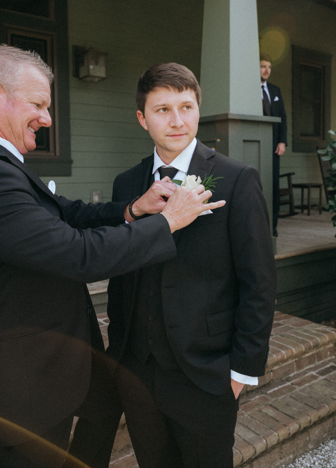 A man in a suit has a boutonniere pinned to his lapel by another man; another person stands in the background on a porch.