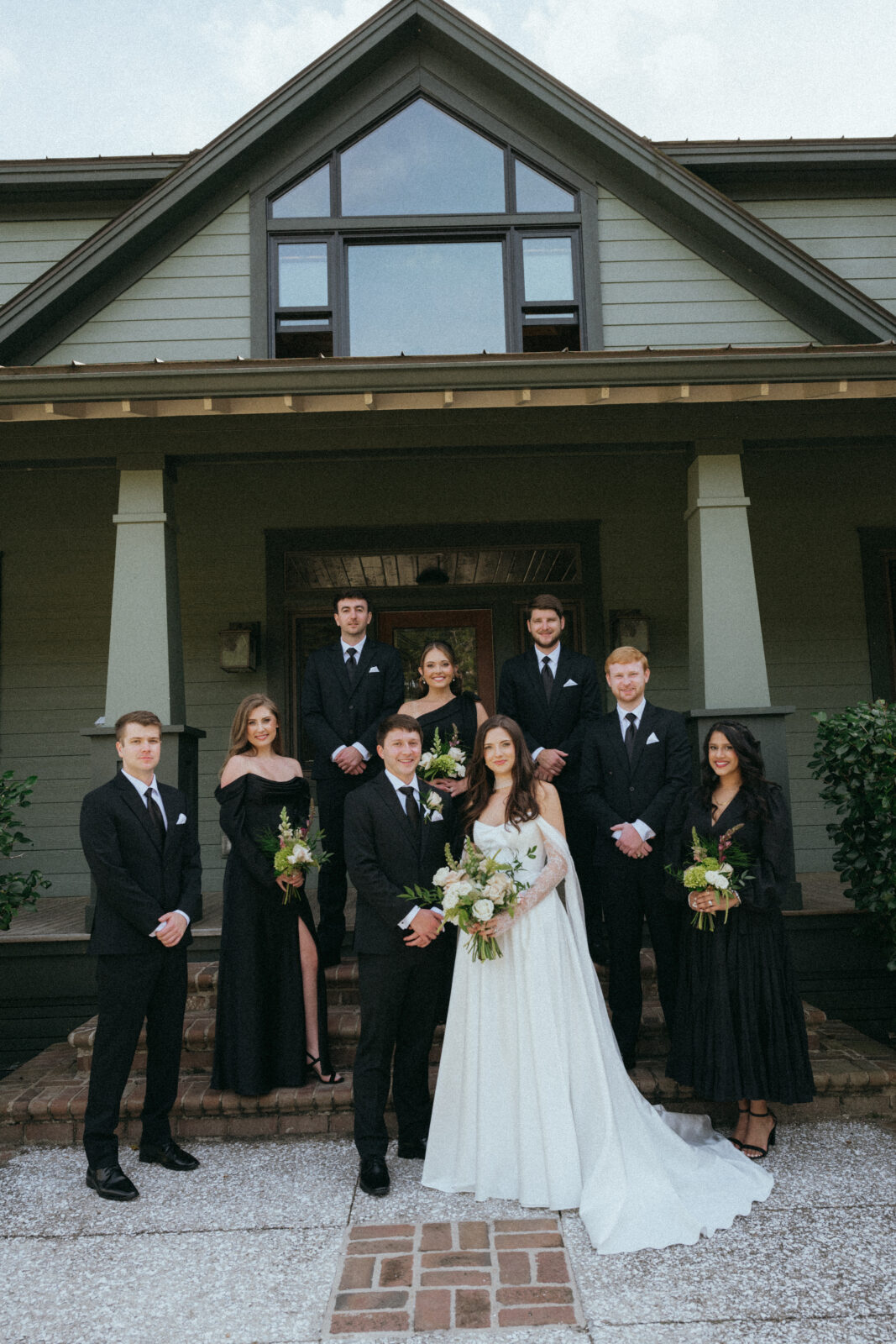 A bridal party of four men in black suits and four women in black dresses poses on the front steps of a sage green house, with the bride in a white wedding gown in the foreground.