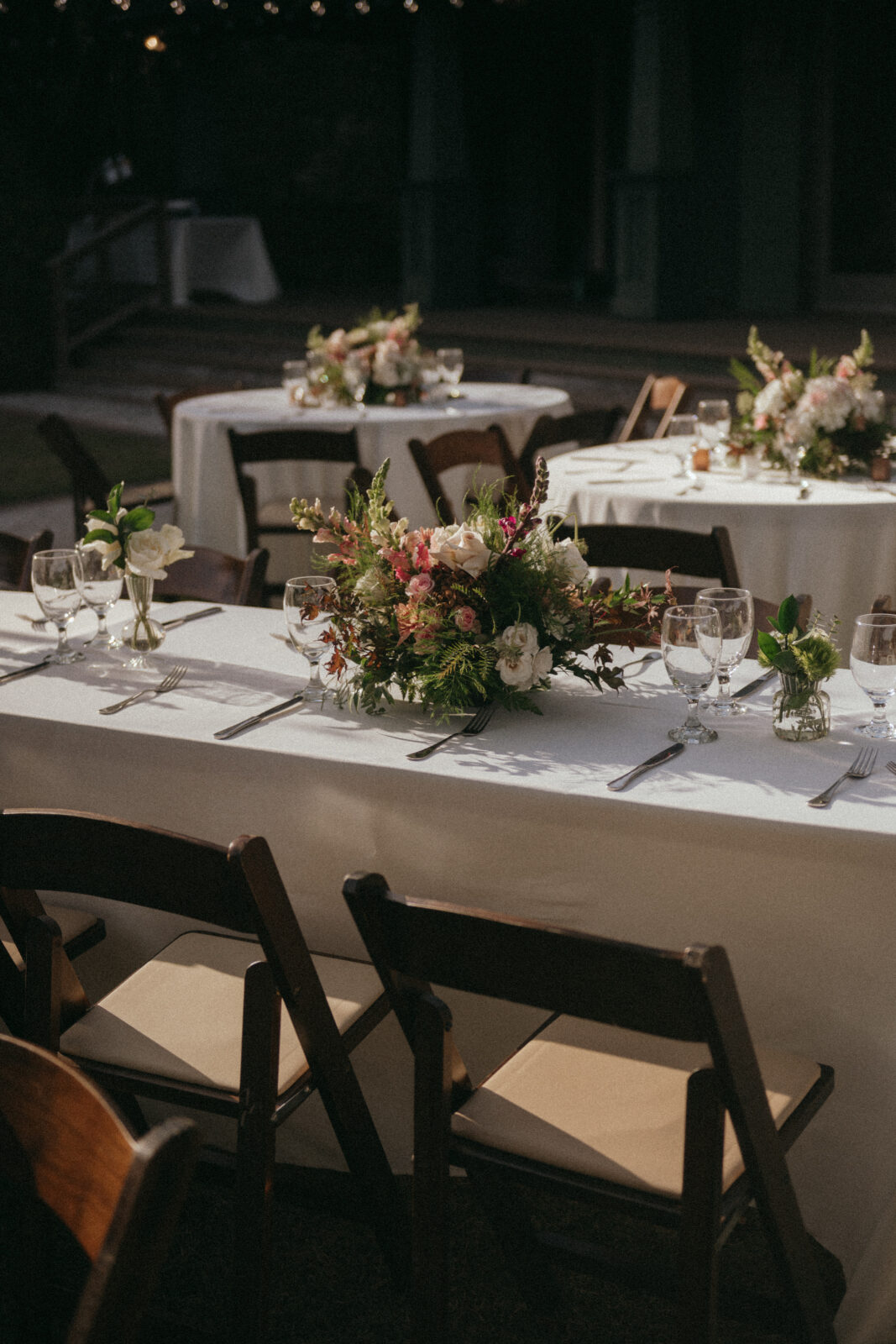 Outdoor tables set with white cloths, floral centerpieces, glassware, and cutlery, surrounded by wooden chairs in natural light.