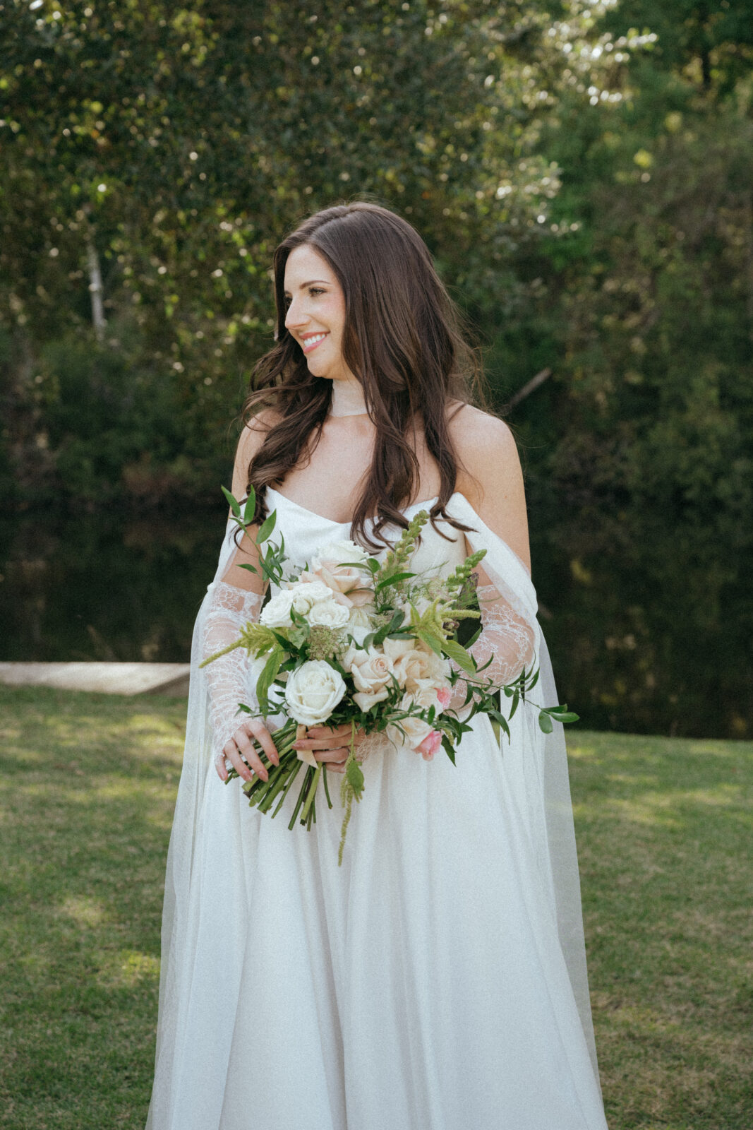 A woman in a white wedding gown and lace arm sleeves holds a bouquet of white and blush roses in a grassy outdoor setting, smiling as she looks to the side.