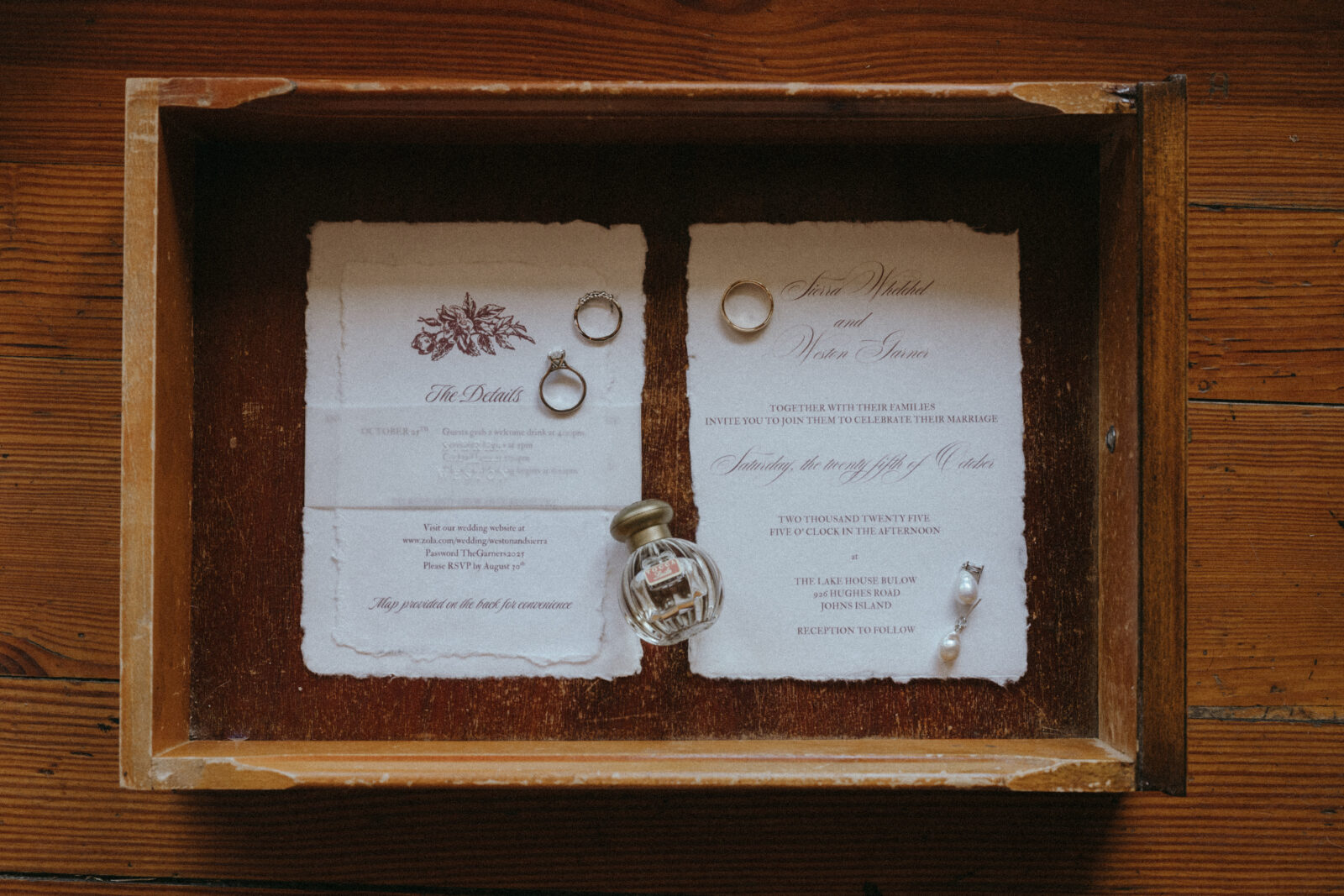 A wooden drawer holds two wedding invitations, rings, a small perfume bottle, and a pearl earring on a wooden surface.