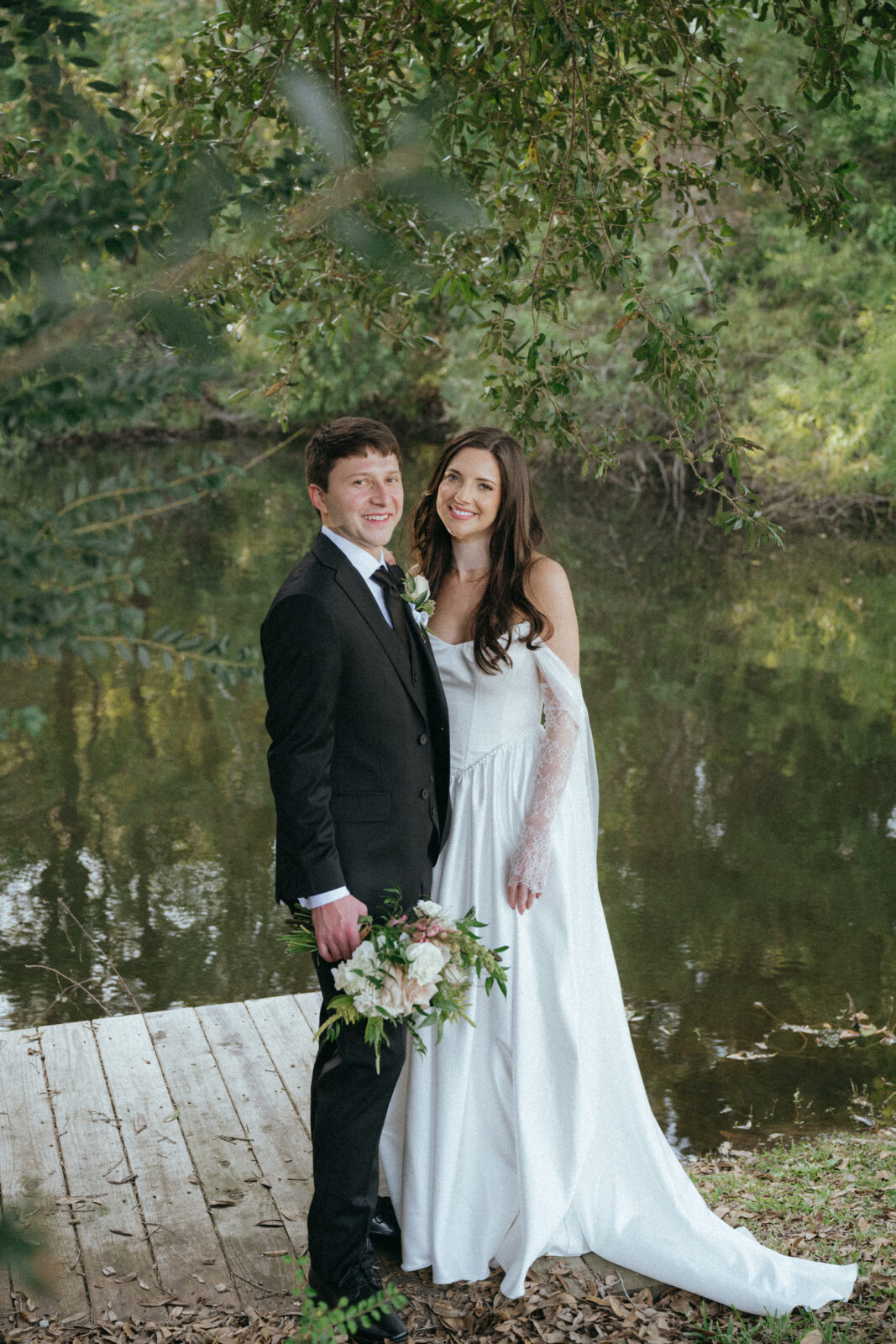 A bride and groom stand together on a wooden dock by a pond, with greenery in the background. The groom holds a bouquet and both are dressed in formal wedding attire, smiling at the camera.