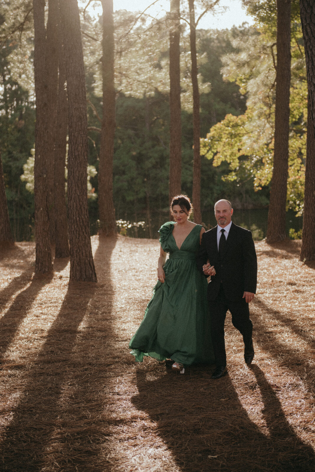 A woman in a green dress and a man in a suit walk together through a sunlit forest with tall trees and long shadows on the ground.