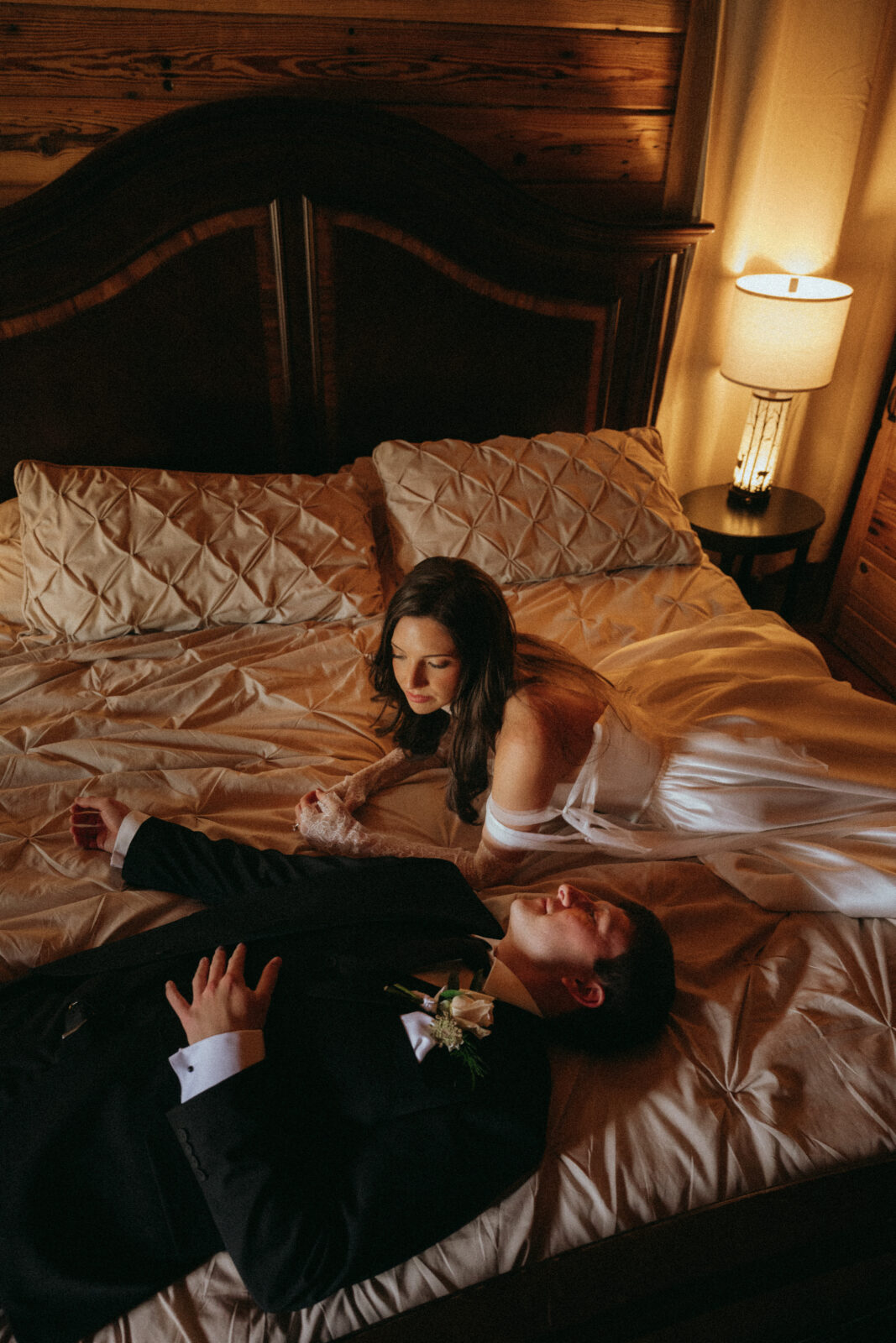 A bride and groom in wedding attire lie side by side on a bed in a warmly lit bedroom, facing each other.