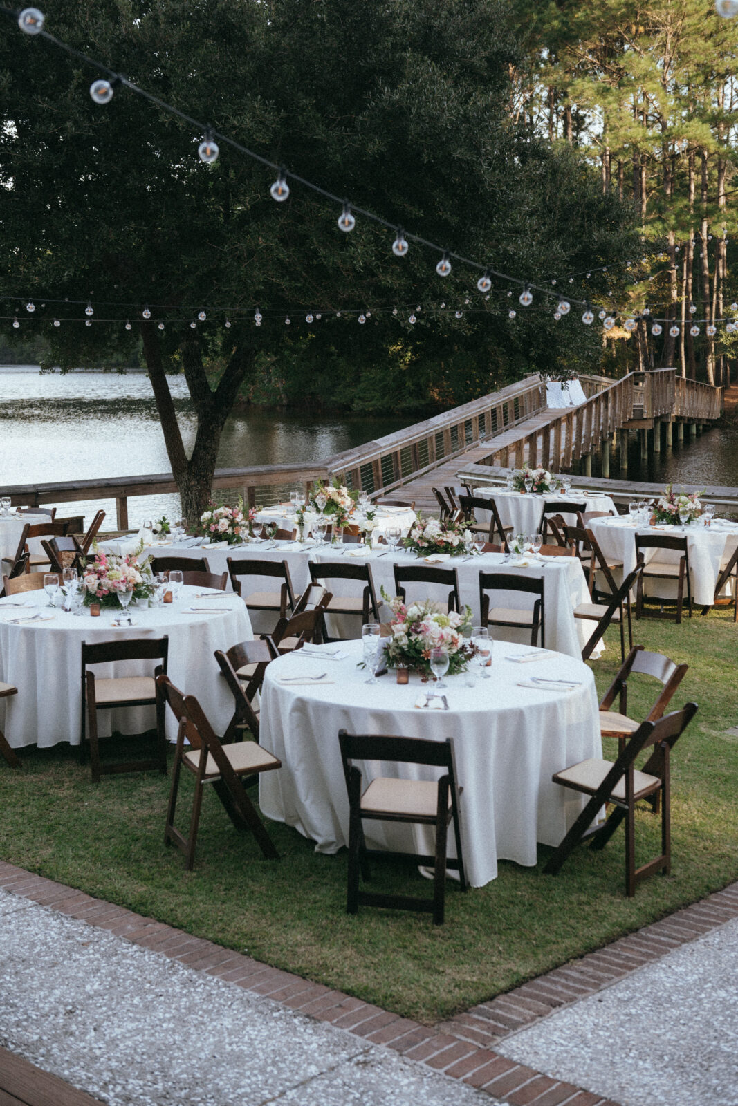 Outdoor event setup with round tables covered in white cloth, floral centerpieces, and chairs, situated by a lake with string lights overhead and a wooden dock in the background.
