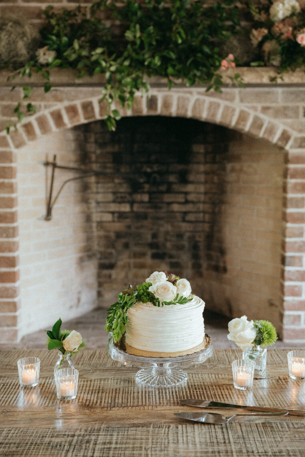 A white frosted cake topped with flowers sits on a glass stand in front of a brick fireplace, with candles and small flower arrangements on the wooden table.