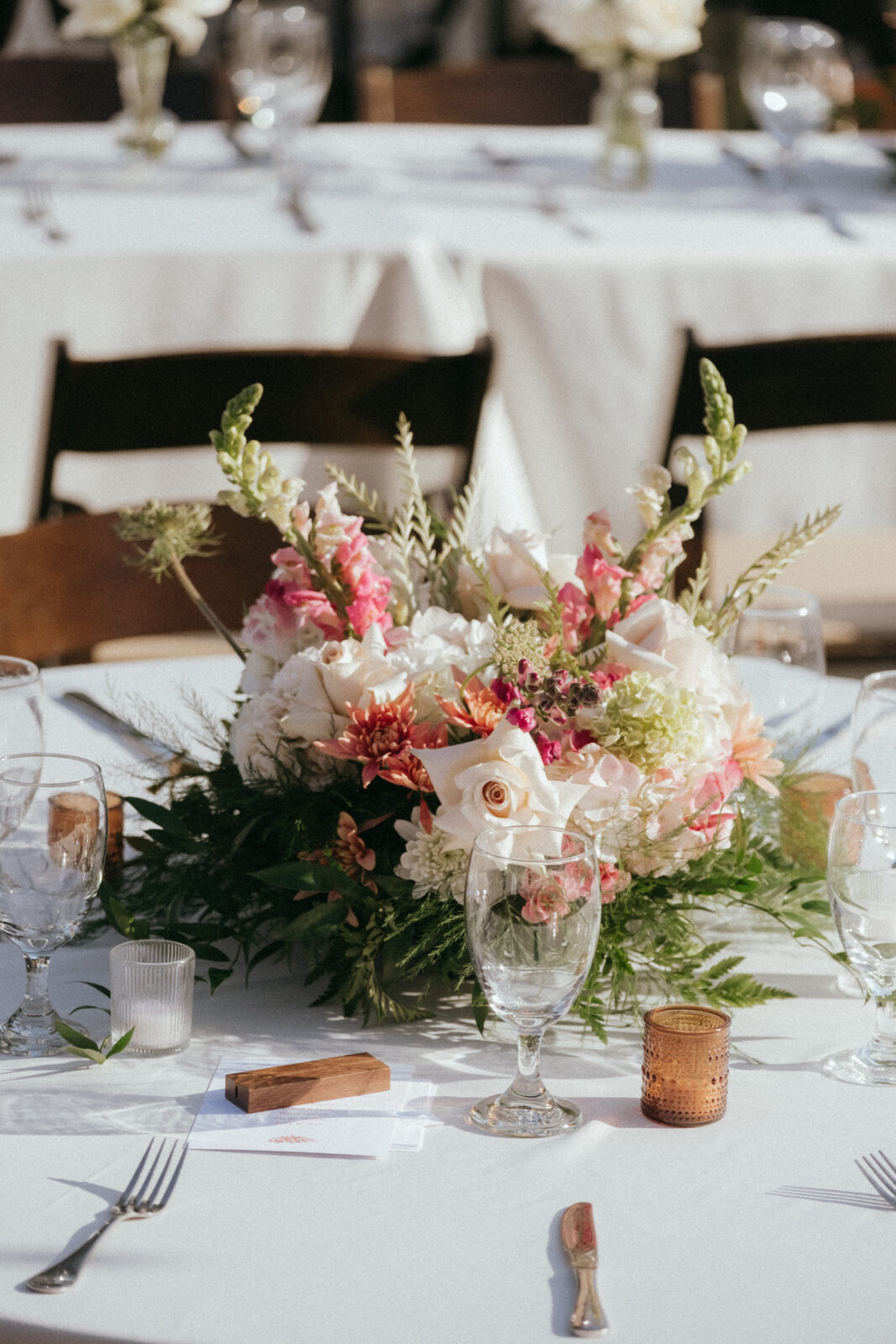 A floral centerpiece with pink and white flowers sits on a white tablecloth, surrounded by glassware, candles, and place settings.