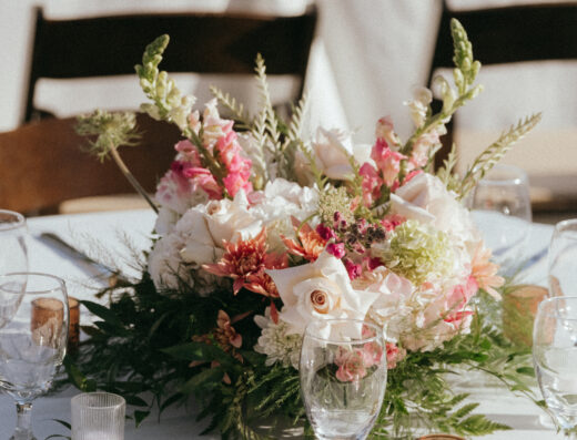 A floral centerpiece with pink and white flowers sits on a white tablecloth, surrounded by glassware, candles, and place settings.