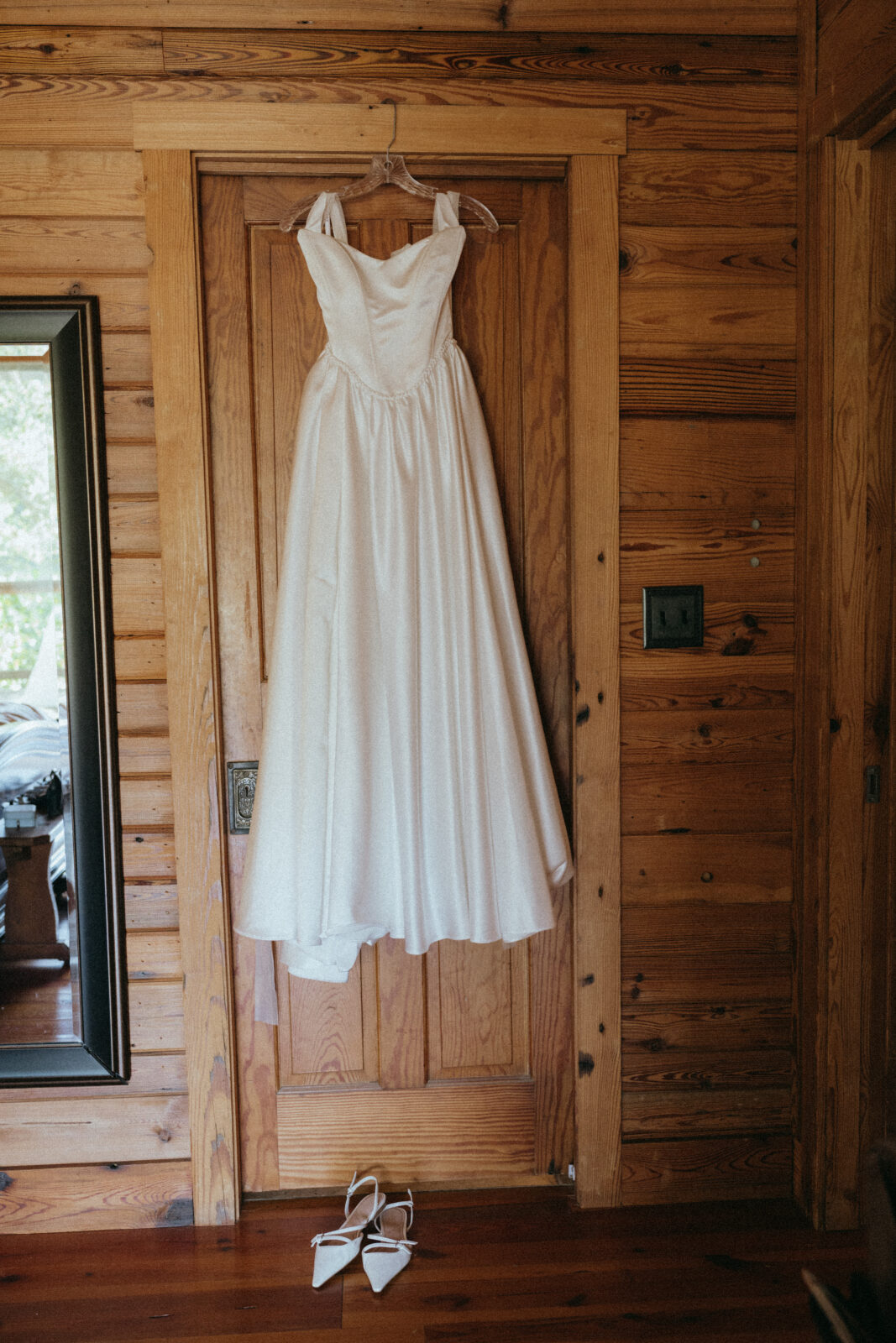A white sleeveless wedding dress hangs on a wooden door, with a pair of white pointed-toe heels placed on the floor below.