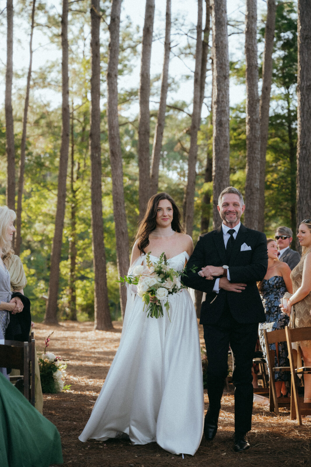 A bride in a white dress holding a bouquet walks down an outdoor aisle on the arm of a man in a black suit, surrounded by seated guests and tall trees.