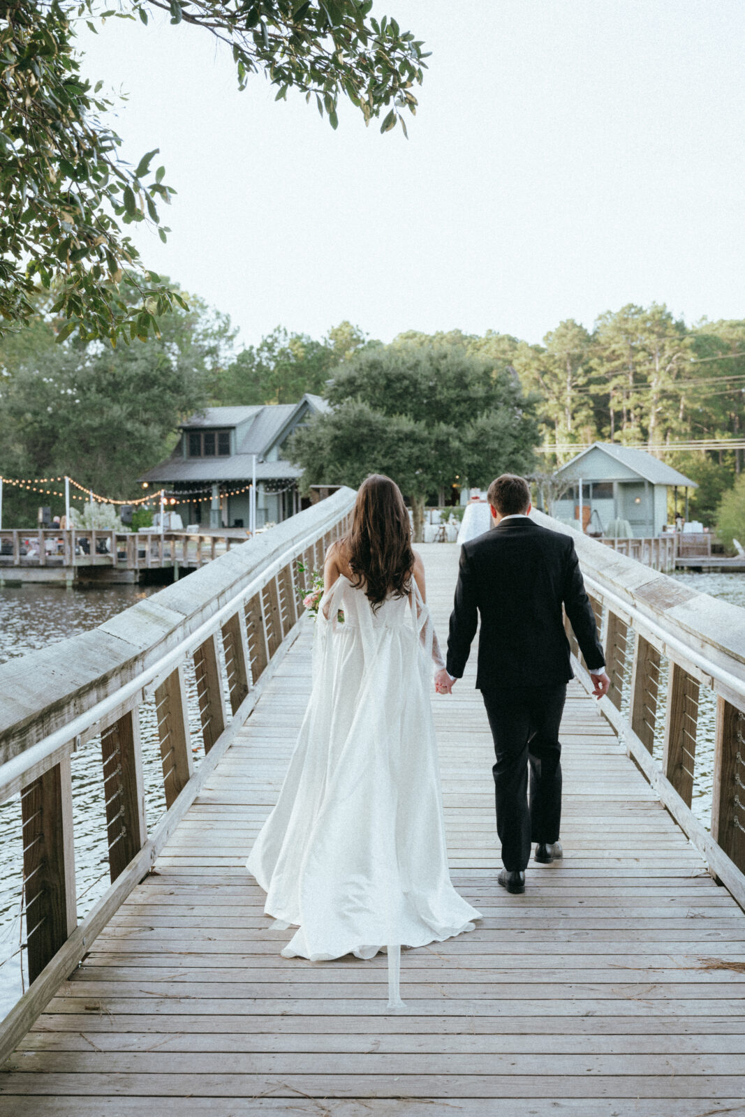 A bride and groom walk hand in hand on a wooden bridge toward lakeside buildings, surrounded by trees.