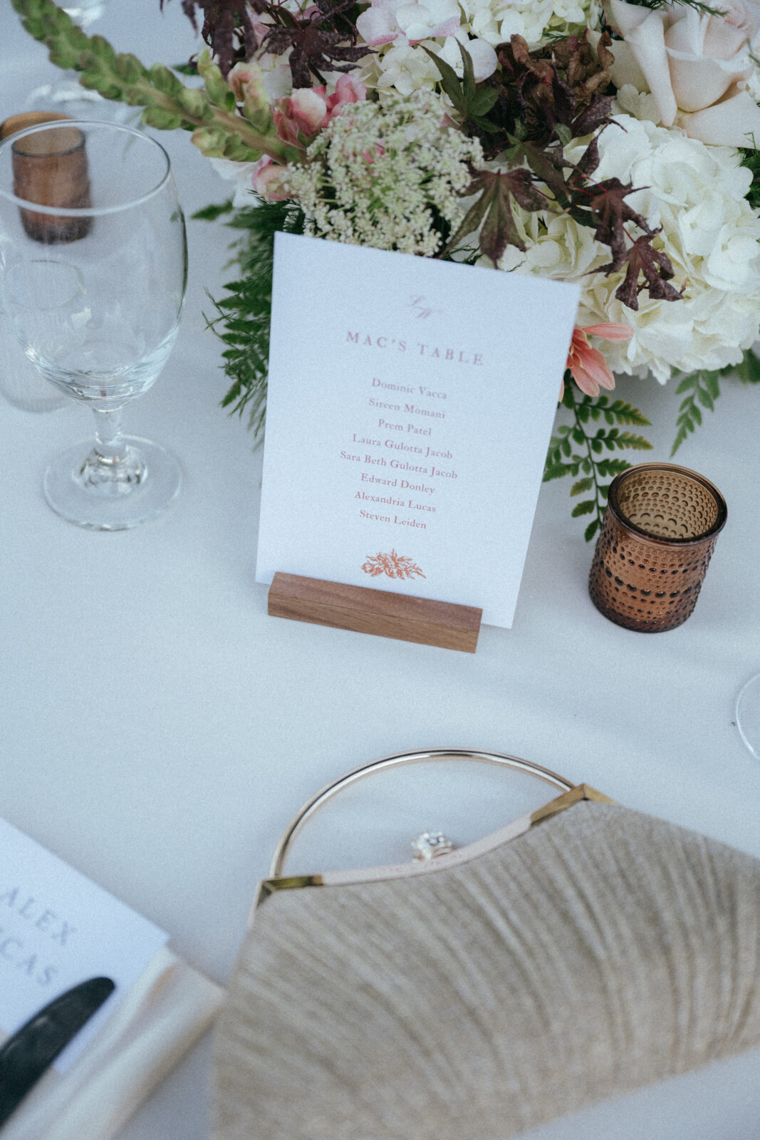 A top-down view of a wedding table setting featuring a floral centerpiece, a glass, a textured clutch, and a sign labeled “Mac’s Table” with a list of guest names.