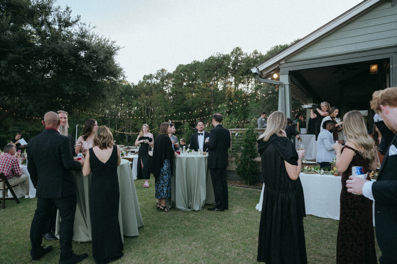 People in formal attire gather outdoors at cocktail tables during an event. String lights hang above, and some guests stand near a house. Trees and greenery are visible in the background.