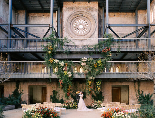A couple kissing under a floral archway in an open-air building with brick walls and metal balconies, facing rows of empty wedding chairs.