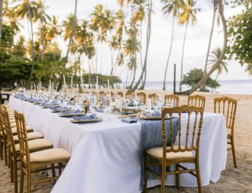 A long, formal dining table set for an event on a sandy beach under palm trees.