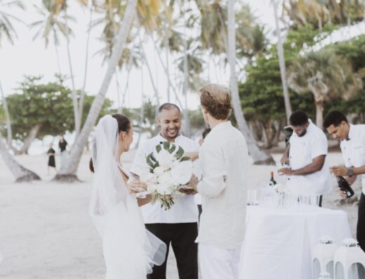 A bride and groom stand on a beach with an officiant as they participate in a wedding ceremony, with staff preparing drinks at a nearby table.