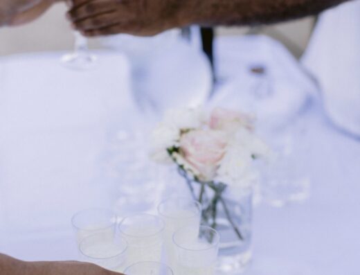 Hands reaching for glasses of sparkling wine on a silver tray, with a vase of flowers and a wine bottle on a white tablecloth.