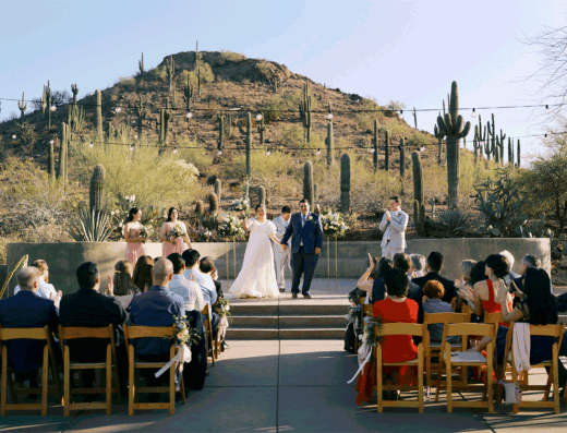 A couple walks down the aisle after their outdoor wedding ceremony in front of a saguaro-covered hillside.