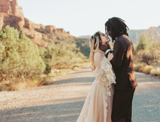 A couple in wedding attire embracing on a desert road with red rock mountains in the background.